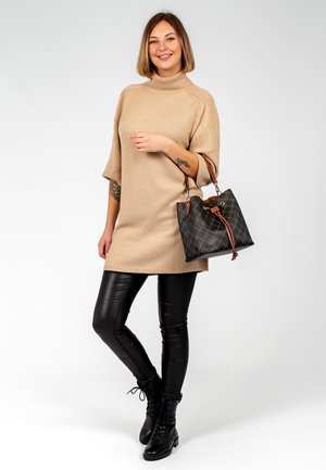 Beige oversized trui jurk met een hoge hals, gecombineerd met zwarte nep leren leggings en zwarte veterschoenen. Houdt een zwarte gequilt handtas vast.