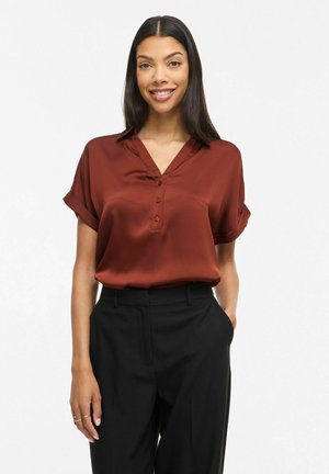 Blouse en satin couleur rouille avec un col en V et une patte de boutonnage. Manches courtes retroussées, texture lisse, portée avec un pantalon noir.