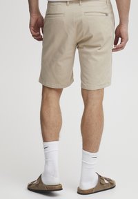 Beige Baumwollshorts mit zwei Gesäßtaschen, flachem Bund und normaler Passform. Weiße Sportsocken und braune Sandalen runden das Outfit ab.