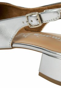 Sandale métallique argentée avec un talon carré, dotée d'une sangle à boucle et d'une semelle intérieure rembourrée. Texture lisse avec une finition brillante.