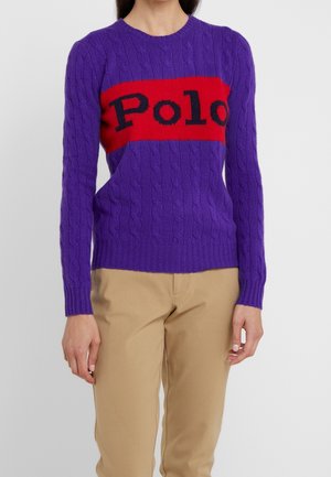 Kobieta ubrana w fioletowy sweter o splecionej fakturze z czerwonym paskiem i czarnym napisem „Polo”, w połączeniu z beżowymi spodniami, na jednolitym tle.