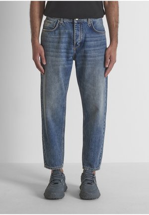 Hombre vestido con vaqueros azules desteñidos, camisa negra y zapatillas grises, de pie frente a un fondo claro y sencillo.