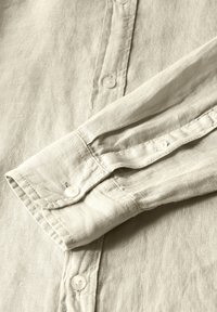 Lněná košile, světle béžové barvy, s límcem na knoflíky a manžetami s perlovo-podobnými knoflíky. Měkká textura, viditelné detaily tkaniny.