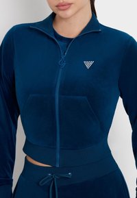 Donna che indossa una giacca blu in velluto con zip e logo triangolare di cristalli, abbinata a pantaloni con cintura coordinata, mostrata dalla testa alle anche.