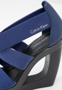 Sandale à talon compensé Calvin Klein présentant un dessus en tissu et cuir bleu, une épaisse semelle noire et deux sangles croisées pour un maintien sécurisé.