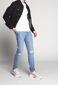 Tmavě modrá džínsová bunda, bílý tričko, světle modré roztrhané džíny a šedé sportovní boty se žlutými akcenty. Důraz na ležérní streetwear styl.