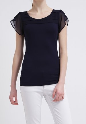 Granatowy top z krótkim rękawem z warstwowymi przezroczystymi akcentami na ramionach. Dopasowany krój, gładka faktura, noszony z białymi spodniami.