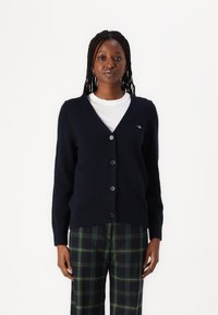 Navy v-neck cardigan with long sleeves, button closure, ribbed hem, and logo detail on the left chest, paired with checkered pants.