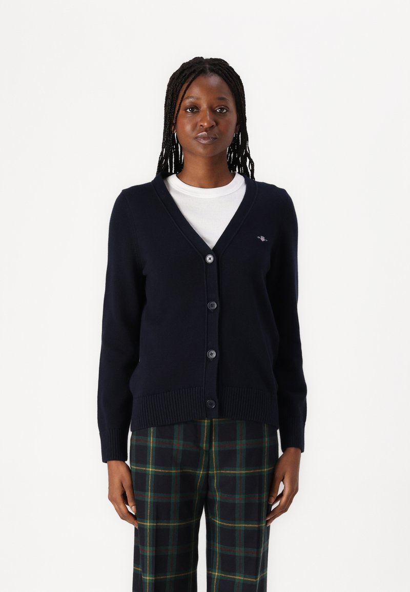 Navy v-neck cardigan with long sleeves, button closure, ribbed hem, and logo detail on the left chest, paired with checkered pants.