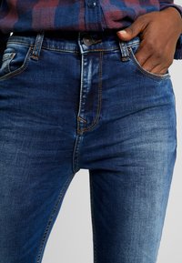 Jeans de mezclilla azul oscuro con una cintura de altura media, costuras visibles y un diseño clásico de cinco bolsillos. Las secciones desgastadas añaden textura.