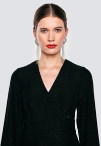 Žena se slicked-back vlasy, nosící dlouhé stříbrné řetězové náušnice, jasně červenou rtěnku a černý top s V-výstřihem na světlém šedém pozadí.