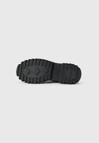 Suela de zapato de goma negra con surcos profundos, patrones en forma de diamante en el centro y un logo circular cerca del talón, sobre un fondo liso.