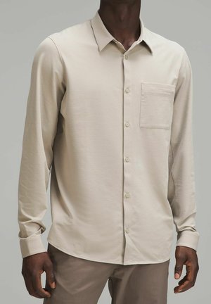 Mężczyzna ubrany w beżową koszulę na długi rękaw z kieszenią na piersi, w połączeniu z jasnobrązowymi spodniami na jednolitym tle.