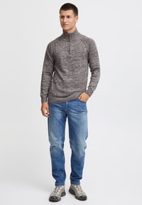 Grå, teksturert zip-up genser med høy krage; kombinert med lyseblå jeans og grå sneakers. Hender i lommene, stående posisjon.