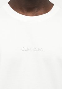 Κοντινό πλάνο ενός ατόμου που φοράει λευκό T-shirt με στρογγυλή λαιμόκοψη και ανάγλυφη επιγραφή "Calvin Klein" στο στήθος.