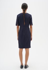 Marinblå klänning med korta ärmar, sidofåror, A-linjeform och sömdetalj bak, bärs med svarta slip-on-sko.