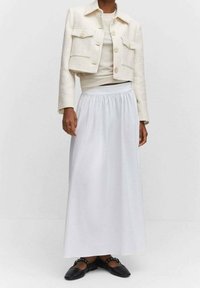 Valkoinen maksihame, jossa on joustava vyötärö, yhdistettynä beigeen teksturoituun lyhytjakkuun, jossa on kaksi etutaskua ja suuret napit. Mustat tasapohjaiset kengät.