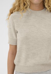 Persoană îmbrăcată cu un pulover scurt din tricot de culoare bej deschis, cu guler, mansete și tiv cu dungi, arătând partea superioară a trunchiului și părul blond, creț.