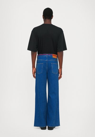 Jeans de mezclilla anchos de color azul con una cintura de media altura, que presentan bolsillos traseros y una cinturilla de color contrastante. Combinados con una camiseta negra de manga corta.