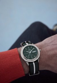 Het Timex Marlin horloge heeft een groene wijzerplaat met dag- en datumweergave, een zilveren roestvrijstalen kast en een groene stoffen band met witte strepen.