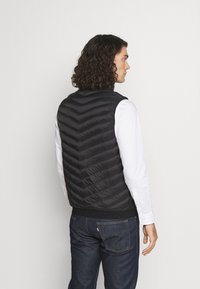 Svart vadderad väst med chevron-quiltat mönster, ribbad nederkant, utan ärmar, bärs över en vit skjorta, med mörka jeans.