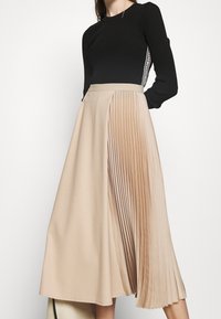 Gonna plissettata beige con un top nero aderente. La gonna presenta una texture liscia su un lato e pieghe verticali sull'altro, accentuando il volume.