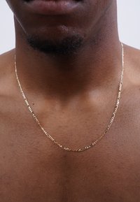 Close-up of bare chest and neck of a person with dark skin wearing a thin gold chain necklace and a small patch of facial hair.