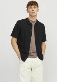 Cardigan negru cu mânecă scurtă și guler, cu nasturi, purtat peste o cămașă bej deschis, asortat cu pantaloni de culoare deschisă. Textură netedă.