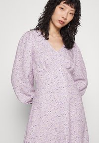 Vestido lilás com mangas bufantes e decote em V, apresentando um padrão floral em roxo e amarelo. Botões ao longo da frente. Textura de tecido suave.