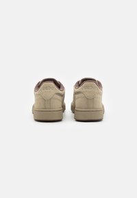 Beige mockasneakers med texturerade beige gummisulor, med en mörkröd klackdetalj och präglad logotyp på baksidan. Rundad form.