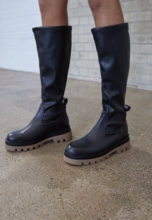 Czarny skórzany botki do kolan z gumową podeszwą. Gruba brązowa podeszwa z wyraźnym wzorem i subtelną teksturą. Wyposażone w uchwyty ułatwiające zakładanie.