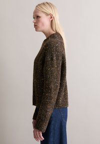 Brązowy, teksturowany sweter z wielokolorowymi kropkami, okrągłym dekoltem i długimi rękawami, w połączeniu z niebieską spódnicą dżinsową. Widok z boku.