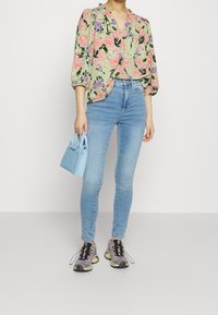 Blouse verte à motif floral avec manches bouffantes, sac à main bleu, jeans skinny bleu clair et chaussures de sport grises avec des accents colorés.