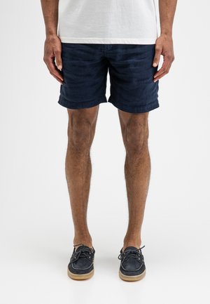 Mann trägt marineblaue legere Shorts, weißes T-Shirt und schwarze Bootsschuhe, steht vor neutralem weißem Hintergrund.
