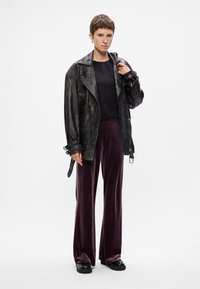 Μαύρο δερμάτινο μπουφάν μηχανής με μεγάλα πέτα, σε συνδυασμό με σκούρες μοβ велούδ παντελόνες με φαρδιές γραμμές. Το μοντέλο κρατά το μπουφάν με το ένα χέρι, φορά μαύρα παπούτσια.