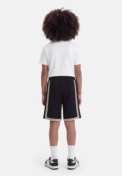 Παιδί με σγουρά μαλλιά που φοράει άσπρο κοντομάνικο μπλουζάκι, μαύρες βερμούδες με διπλές μπεζ πλαϊνές ρίγες, λευκές κάλτσες και μαύρα-άσπρα αθλητικά παπούτσια, όρθιο.