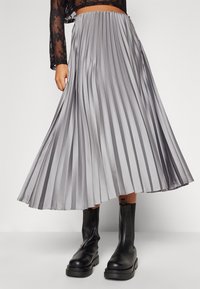 Plisovaná maxi sukně ze saténové látky, stříbrné barvy, s hladkým povrchem a širokým pasem, kombinovaná s masivními černými kotníkovými botami.