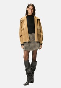 Manteau oversize en laine beige avec grandes poches, pull noir, mini-jupe à carreaux et bottes en cuir noires hauteur genou avec détail plissé.