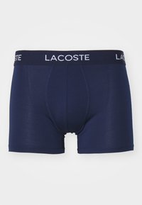 Sötétkék boxeralsó, testhezálló fazonban, fekete derékrésszel, amelyen fehér betűkkel a "LACOSTE" felirat található, sima textúrával és minta nélkül.