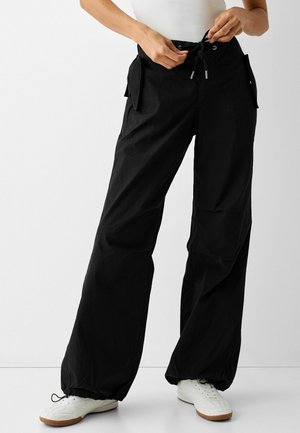 Zwarte, lichte, losse broek met een elastische tailleband met trekkoord, zijzakken en elastiek bij de enkels, gecombineerd met witte schoenen.