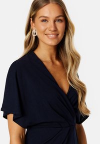 Námořnické zavinovací šaty s krátkými, splývavými rukávy a výstřihem do V; obsahují nařasené detaily v pase a mají hladkou, jemnou texturu látky.