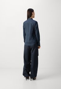Camicia blu navy a maniche lunghe con colletto, abbinata a pantaloni larghi di un blu navy scuro, mostrata da una prospettiva posteriore.