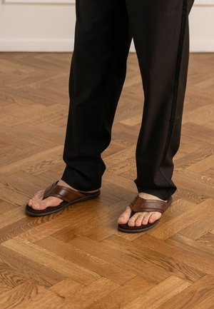 Voeten in bruine leren sandalen met zwarte zolen, gedragen met zwarte broek op een houten parketvloer.