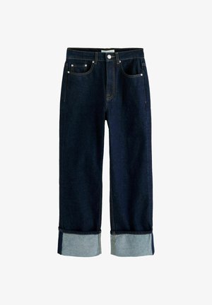 Tumši zili džinsa džinsi ar platiem kāju galiem, atlocītām malām, piecām kabatām un redzamu dzeltenu diegu uz balta fona.