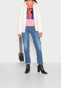 Witte corduroy jas, grafische roze en oranje trui met leeuwenontwerp, blauwe jeans met rechte pijpen, zwarte enkellaarsjes met hak, zwarte handtas.