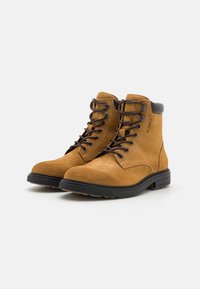 Bottines en daim beige avec un bout arrondi, un design à lacets et une semelle en caoutchouc noire. Présente des accents marron au niveau du col et des œillets.