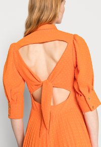 Vestido laranja com um tecido texturizado, apresentando mangas bufantes, um decote em forma de buraco com um laço nas costas e detalhes de saia plissada.