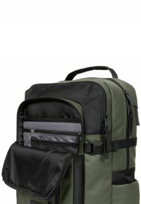 Mochila verde con acentos negros, que presenta múltiples compartimentos con cremallera, bolsillos de malla y un robusto asa superior. Exterior texturizado.
