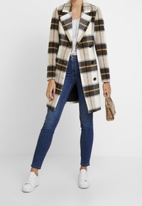 Romige geruite jas met zwarte en oranje strepen, dubbelzijdig ontwerp, gecombineerd met donkerblauwe skinny jeans en witte sneakers.