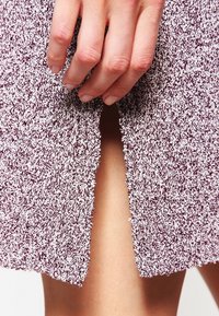 Texturerad lila stickad klänning med en sidofåra, med en hand som försiktigt greppar tyget. Närbild som visar material och detaljer.